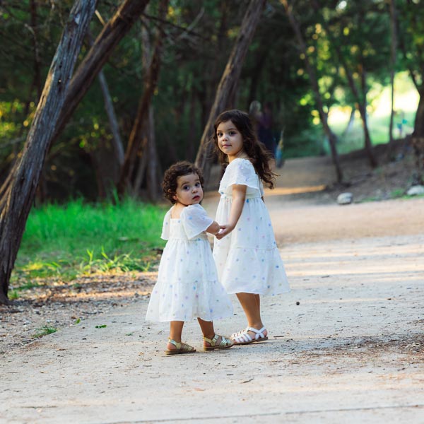Two young girls in white dresses holding hands on a forest trail in Dripping Springs, TX