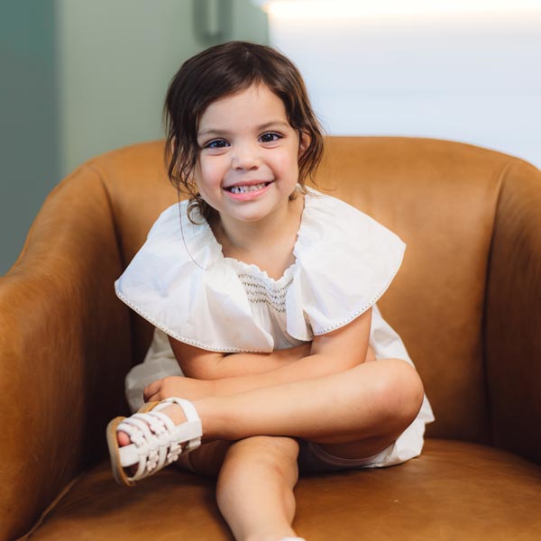 Smiling young girl in a white dress sitting on a brown chair in Dripping Springs, TX